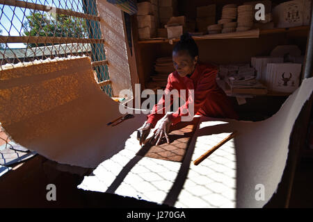 Una donna di taglio di un foglio di carta fatta a mano in carta-making facility in Ambalavao Madagascar; prodotti cartacei finiti su un ripiano dietro di lei Foto Stock