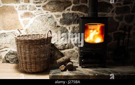 Il focolare e camino all'interno di una parete di pietra cottage con una stufa a legna, registri e cesto in un ambiente accogliente e famigliare scena. Foto Stock