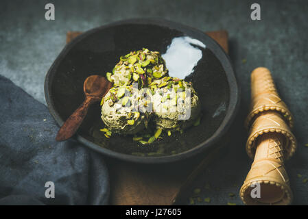 In casa il pistacchio ice cream scoop con si è schiantato i pistacchi in piastra scuro su tavola di legno, coni di cialda su grigio sfondo concreto, selettivo Foto Stock