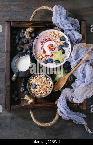 Ciotola frullato salutare prima colazione. Yogurt alla fragola con mirtilli, granola, menta, brocca del latte e carambole nel vassoio in legno sui prodotti tessili tessuti a punto di garza oltre dar Foto Stock