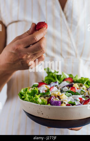 Una donna in possesso di una ciotola di fragole fresche insalata di pasta una spruzzata di yogurt in una mano e afferrando una fragola con l'altra. Foto Stock