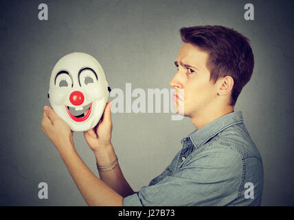 Giovane uomo triste con happy clown maschera isolata sul muro grigio Sfondo. Le emozioni umane. Personalità scissa concept Foto Stock