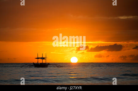 Barca a vela al tramonto con fantasia a profilarsi in barca a vela lungo contro una vivida tramonto colorato di arancione e giallo pieno di colore del cielo. Foto Stock