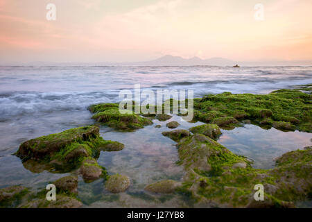 Puntello roccioso coperto con alghe verdi con lo splendido oceano nelle prime ore del mattino con vedute del vulcano e delle montagne. Indonesia Bali Foto Stock