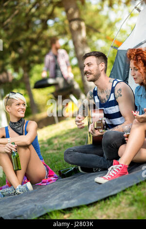 Guy suonare la chitarra e le ragazze gode nella parte anteriore della tenda al Summer Camp in foresta Foto Stock