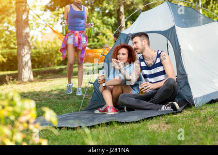 Tatuato guy e lassie si siede e beve birra nella parte anteriore della tenda al campo estivo in natura Foto Stock