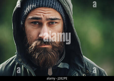 Outdor porttrait di un bel brutale uomo barbuto con scuri di barba e baffi vestito in abiti invernali, coperto di brina. Uomo di rivestimento antiusura hat e ho Foto Stock