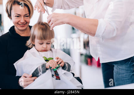 Sezione mediana del barbiere il taglio ai capelli della ragazza seduta con la madre in salone Foto Stock