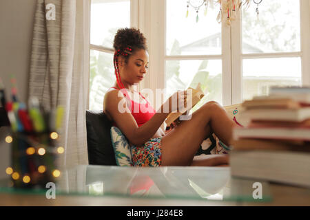 Felice studentessa la lettura del libro e seduto sul divano. Giovane africano donna americana rilassante, nero ragazza sdraiato sul lettino. Persone di origine ispanica e stile di vita Foto Stock