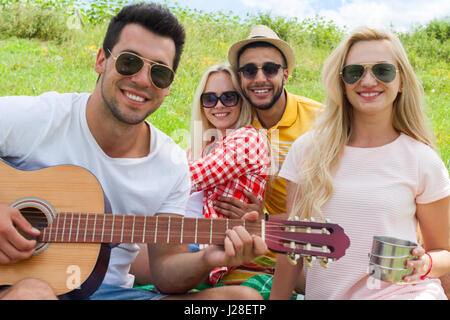 I giovani in ascolto ragazzo a suonare la chitarra gruppo amici estate il giorno seduto erba verde picnic esterno natura accoppiare due uomini con le ragazze ridono cantando s Foto Stock