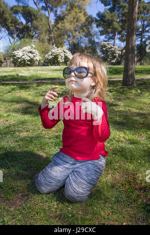 Divertente ritratto di blonde bimbo di tre anni, con maglietta rossa, seduta in erba verde nel parco, giocando con oversize donna adulta occhiali neri Foto Stock