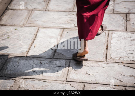 I piedi di un monaco buddista indossa sandali e il tradizionale abito rosso camminando nel sole di mezzogiorno con un fortemente definito ombra. Il Monastero di Kopan, Kathmandu, Foto Stock