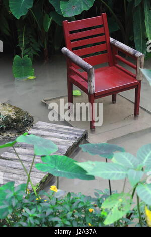 Maroon di legno verniciato poltrona posizionato nel mezzo di acqua e collegata a terra attraverso una pista di log che serve come pesce spa. Parco delle Farfalle in co Foto Stock