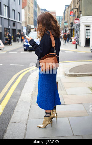 Londra - febbraio, 2017: donna che indossa blu mantello di lana, calze e oro stivaletti in piedi in strada usando il telefono durante la London Fashion Week, verticale Foto Stock