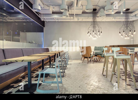 Hall in stile loft in un cafe con pareti luminose e piastrelle bianche e nere sul pavimento. Ci sono divani, diverse sedie, poltrone, rotonda e quadrata Foto Stock