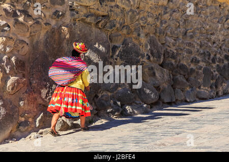 Donna Peruviana cholita vestito nel tradizionale tessuto colorato, che trasportano il sacco e a piedi fino alla strada con mura pietrose, Inkan Valle Sacra, Ollan Foto Stock