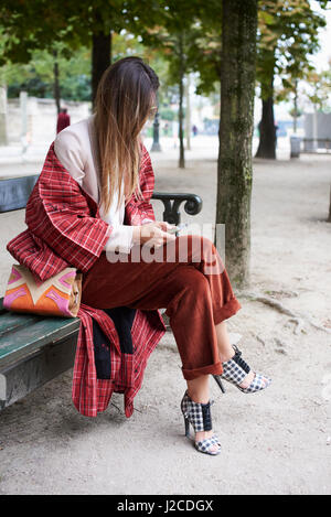 La donna si siede su una panchina nel parco usando il telefono a piena lunghezza verticale Foto Stock