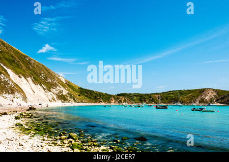 Piccole imbarcazioni da diporto galleggiante sul mare blu a Lulworth Cove lontano dalle rocce la bordatura della spiaggia di ciottoli su cui le persone stanno godendo il sole Foto Stock