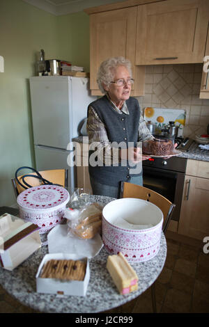 Signora anziana preparare un tè inglese con vari tipi di torte sul suo tavolo da cucina, England, Regno Unito Foto Stock