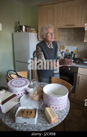 Signora anziana preparare un tè inglese con vari tipi di torte sul suo tavolo da cucina, England, Regno Unito Foto Stock