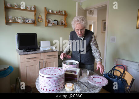 Signora anziana preparare un tè inglese con vari tipi di torte sul suo tavolo da cucina, England, Regno Unito Foto Stock