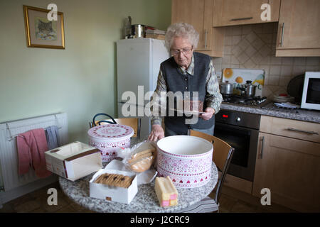 Signora anziana preparare un tè inglese con vari tipi di torte sul suo tavolo da cucina, England, Regno Unito Foto Stock