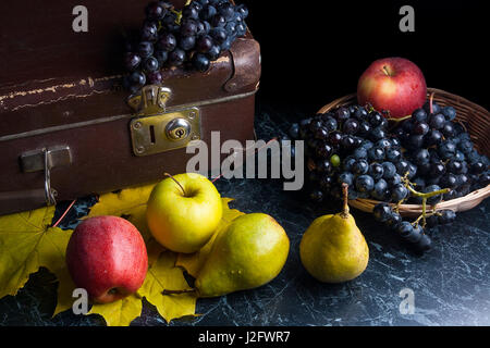 Frutte mature mele, pere e diversi giallo Foglie di autunno sul marmo scuro dello sfondo. L'uva matura nel cestello giallo e il vecchio caso sul retro dello sfondo. Foto Stock