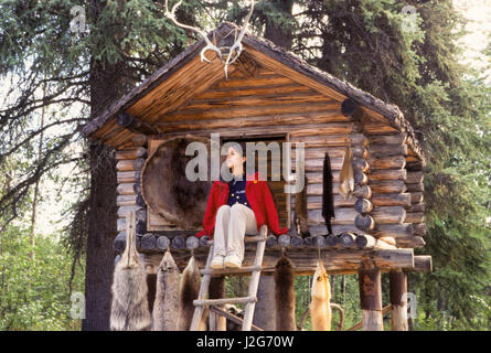 Teenage ragazza Athabaskan siede sulla piattaforma di un registro tradizionalmente costruite nella conservazione di alimenti shelter, chiamata cache, con pelli di animali e una stirata beaver pelle. Situato all'interno di un caccia e pesca camp. Villaggio Chena, Alaska Foto Stock