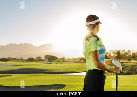 Vista posteriore della giovane donna in piedi sul campo da golf in una giornata di sole. Professional Golfer femmina holding club di golf sul campo e guardando lontano. Foto Stock