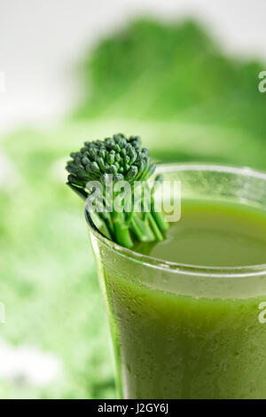 Un frullato verde servito in un bicchiere e rabboccato con uno stelo di broccolini e alcune foglie di cavolo e kale in background Foto Stock