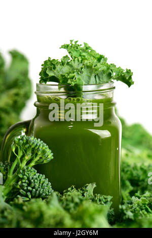 Un frullato verde servito in un bicchiere mason jar circondato da alcune foglie di cavolo e alcuni steli di broccolini su sfondo bianco Foto Stock