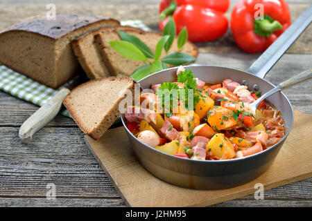 Hot Potato gulasch con pancetta e salsicce di Vienna è servita in una padella di ferro con deliziosi scuro agriturismo cotto il pane su un vecchio tavolo in legno Foto Stock