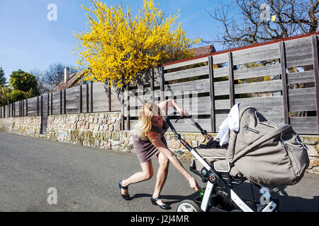 Mamma sulla passeggiata Baby Stroller Cart donna che spinge la carrozza che cammina Pram camminando donna in fretta passeggiata Primavera giornata soleggiata Forsythia Plant Fence tempo libero Foto Stock