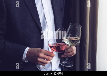 Chiudere orizzontale di uomo caucasico in abito nero e una camicia bianca con due bicchieri alti con rose e vino bianco in un evento dalla finestra di natura Foto Stock