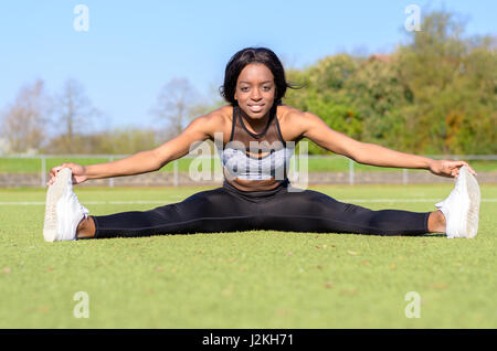 Montare amichevole supple giovane donna facendo i gruppi come ella siede su un verde campo sportivo sorridente felicemente con la videocamera in uno stile di vita attivo concept Foto Stock