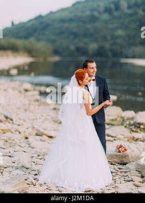 Bella coppia di nozze abbraccia dolcemente vicino alla riva di un fiume di montagna con pietre Foto Stock