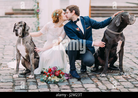 Happy sposi seduti sulle loro haunches e baciare tenendo i loro cani nel centro antico Foto Stock