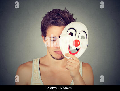 Giovane uomo triste decollare clown maschera isolata sul muro grigio Sfondo. Le emozioni umane Foto Stock