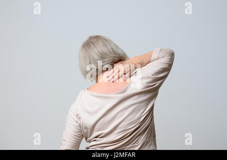 Senior grey-haired donna da lei indietro toccando il collo, come se soffrono di dolore chondrosis, in piedi sulla pianura sfondo grigio Foto Stock