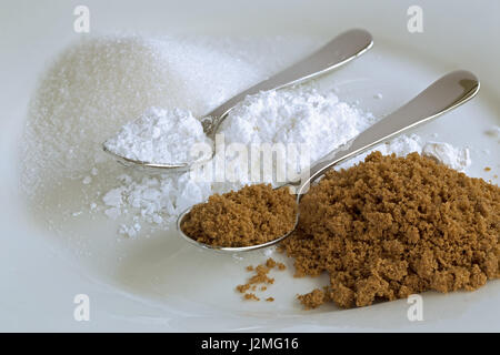 Due cucchiai di lucido su piccoli mucchi di marrone, ghiaccio e lo zucchero cristallizzato su una piastra bianca. Foto Stock
