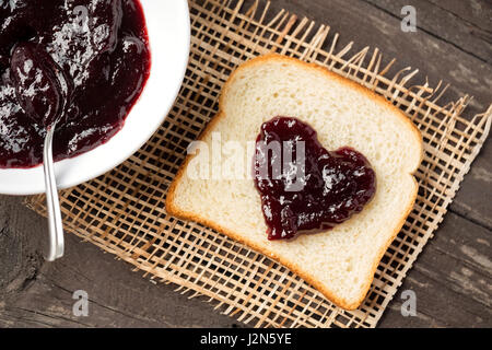 A forma di cuore con pane e marmellata di ciliege su sfondo di legno Foto Stock