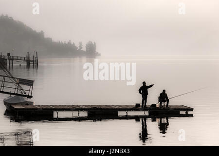 Seduta di pescatori sul molo di pesca con un altro uomo si fermò puntando su un inizio di foschia mattutina a Lenno Lago di Como Italia nel mese di aprile Foto Stock
