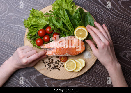 Materie filetti di salmone sul tagliere in legno. Foglie di lattuga, spezie, le fette di limone su una tavola di legno. Woody background. vista dall'alto. Le mani di una donna in Foto Stock