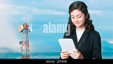 Attraente bussiness donna utilizzando computer tablet con torre di telecomunicazioni contro la gamma della montagna durante il tramonto in background Foto Stock