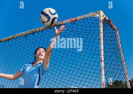 Giovane ragazza asiatica portiere cattura la sfera durante un divertente pratica in un campo su un cielo blu chiaro giorno Foto Stock