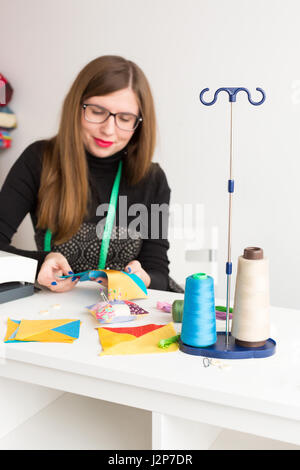 Ricamo e la trapuntatura in officina di una giovane donna di un sarto - sarto sorridente in bicchieri a lavorare con pezzi di stoffa colorata sul tavolo con fili, tessuti, aghi Foto Stock