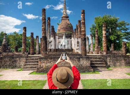 Tourist donna in costume rosso di scattare una foto del Buddha antico con il suo smartphone in Wat Sa Si di Sukhothai Historical Park, Thailandia Foto Stock