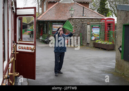 Protezione prezzo Jim soffia il fischietto e solleva una bandiera verde come egli si prepara a bordo di una carreggiata stretta treno a vapore a Woody Bay station sulla riapertura del tratto ferroviario tra Lynton e Barnstaple, Devon. Foto Stock