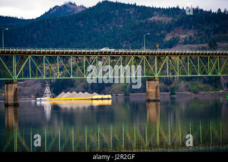 Giallo laden Barge, che spinge il sollevamento, nuota in metallo verde ponte stradale sul fiume, che si riflette nell'acqua su uno sfondo Foto Stock