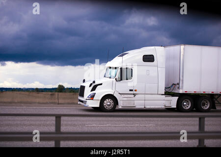 Moderno e confortevole lusso white big rig semi camion con rimorchio in autostrada multilinea con la scherma contro un buio cielo tempestoso Foto Stock